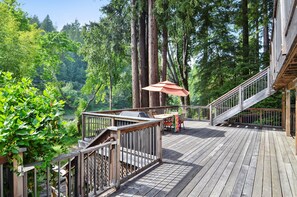 A wooden deck with a grill and outdoor furniture, surrounded by tall trees. - Boathouse ~ On River, Deck, Steps to Beach, Family  Dog Friendly! (Guerneville)