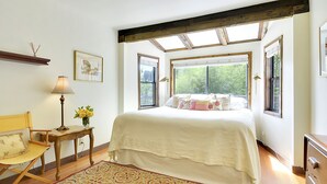 A bedroom with a large bed, a chair, a small table with a lamp, and a window with a view of trees. - Boathouse ~ On River, Deck, Steps to Beach, Family  Dog Friendly! (Guerneville)