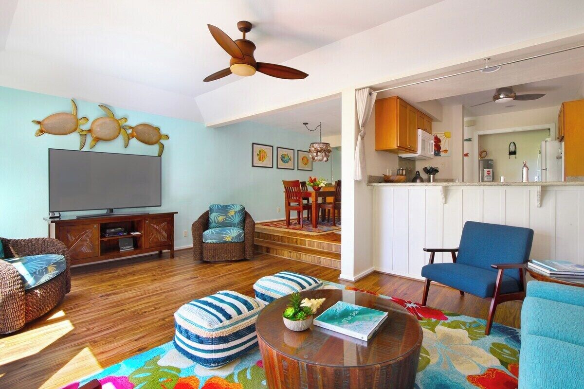 A living room with a TV, seating, a ceiling fan, and a kitchen area visible through an open door.