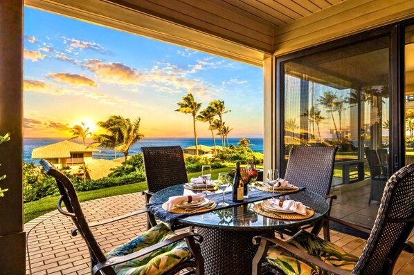 A round glass table with chairs, set for dining, overlooking a scenic view of the ocean and palm trees at sunset.