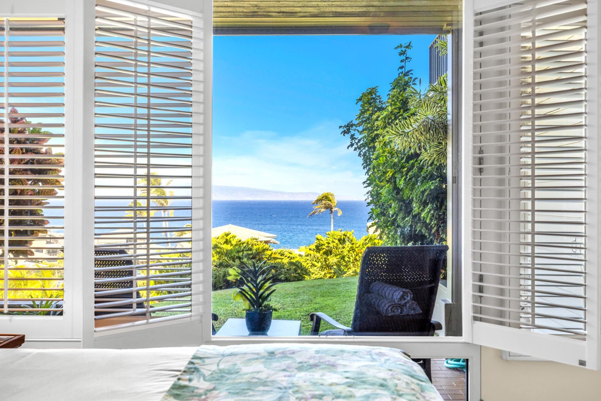 A room with a view of the ocean through open blinds, a potted plant, and a chair.