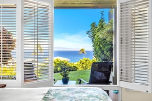 A room with a view of the ocean through open blinds, a potted plant, and a chair.