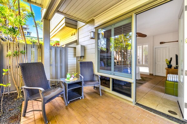 A covered patio with two wicker chairs and a small table, a sliding glass door leading to an interior room, and a wooden fence with plants.