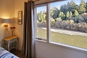 Una stanza con una grande finestra che si affaccia su un fiume e una foresta. C'è un comodino con una lampada e un vaso di fiori.