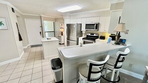 Cozinha moderna com ilha central, armários brancos, eletrodomésticos de aço inoxidável e um pequeno espaço de jantar com duas cadeiras.