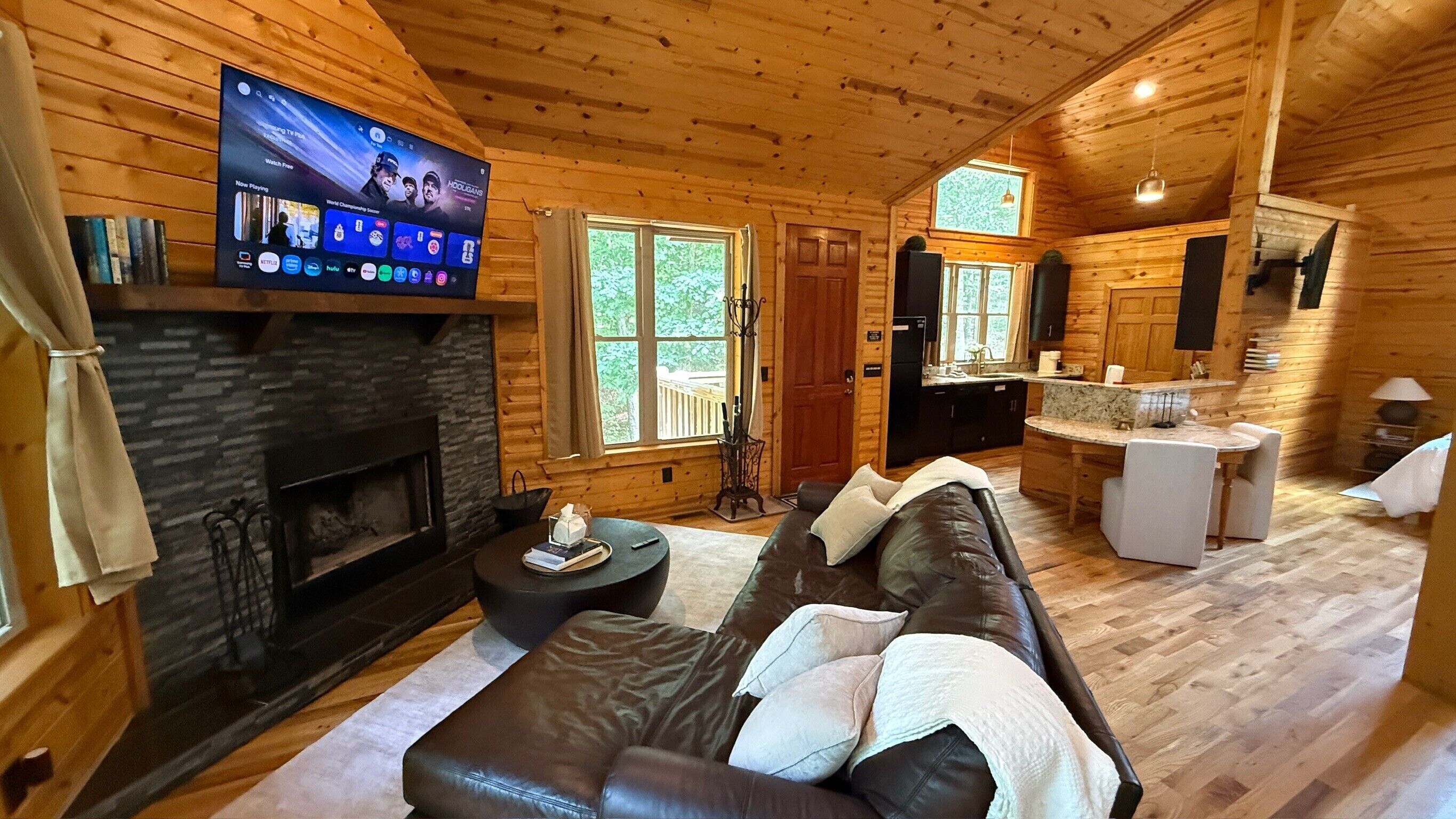 Interior acogedor de una cabaña con chimenea, sofá de cuero, comedor y televisor montado en la pared.