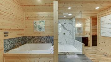Interior de una cabaña de madera con una bañera grande, zona de ducha con paredes de mármol y un pequeño rincón de cocina.