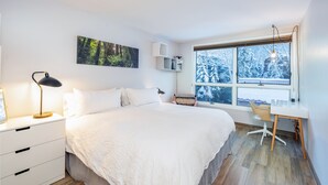 A bedroom with a bed, a desk, a chair, a nightstand, a lamp, a shelf with books, a framed picture on the wall, and a window with a view of trees. - One block walk to gondola, ski lockers downstairs, River Trail close by! (Telluride)