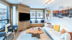 A modern living room with a flat-screen TV, wooden accents, a white sofa, and a large window with a view of a snowy landscape. - One block walk to gondola, ski lockers downstairs, River Trail close by! (Telluride)