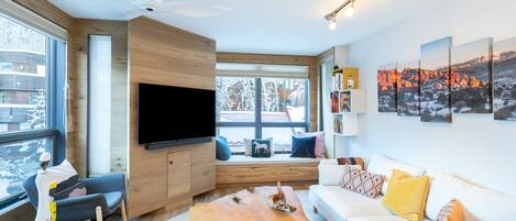 Una sala de estar moderna con un televisor de pantalla plana, detalles de madera, un sofá blanco y un ventanal con vistas a un paisaje nevado.