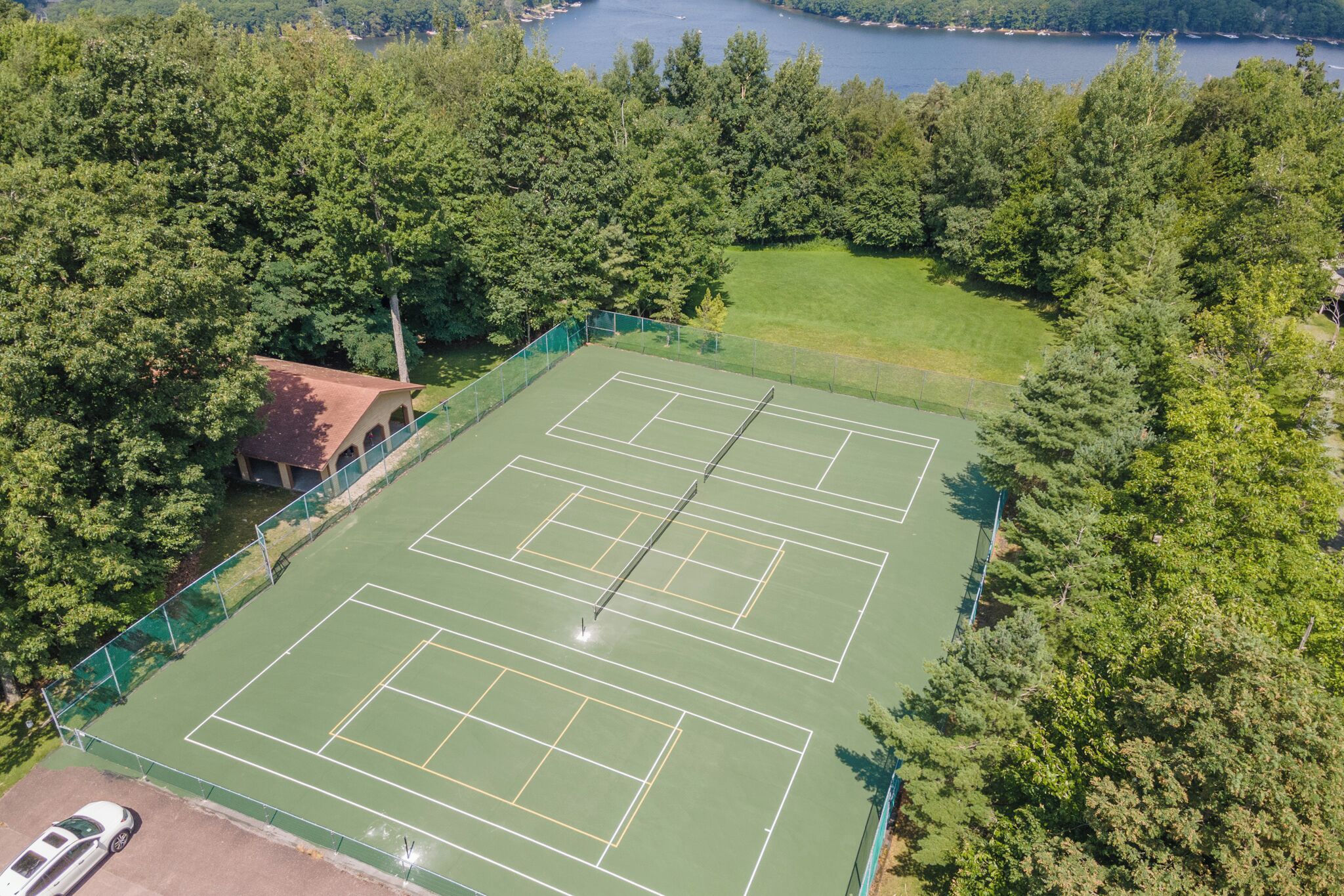 Vista aérea de una cancha de tenis rodeada de árboles frondosos, con un pequeño edificio a la izquierda.