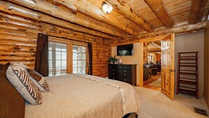 A log cabin bedroom with a bed, dresser, TV, and a door leading to another room. - Mountain Escape w/ Fire Pit, Pool Table & Hot Tub! (OAKLAND)