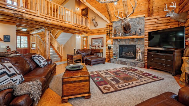 A spacious log cabin interior with a fireplace, leather furniture, a staircase, and a mounted deer head. - Mountain Escape w/ Fire Pit, Pool Table & Hot Tub! (OAKLAND)
