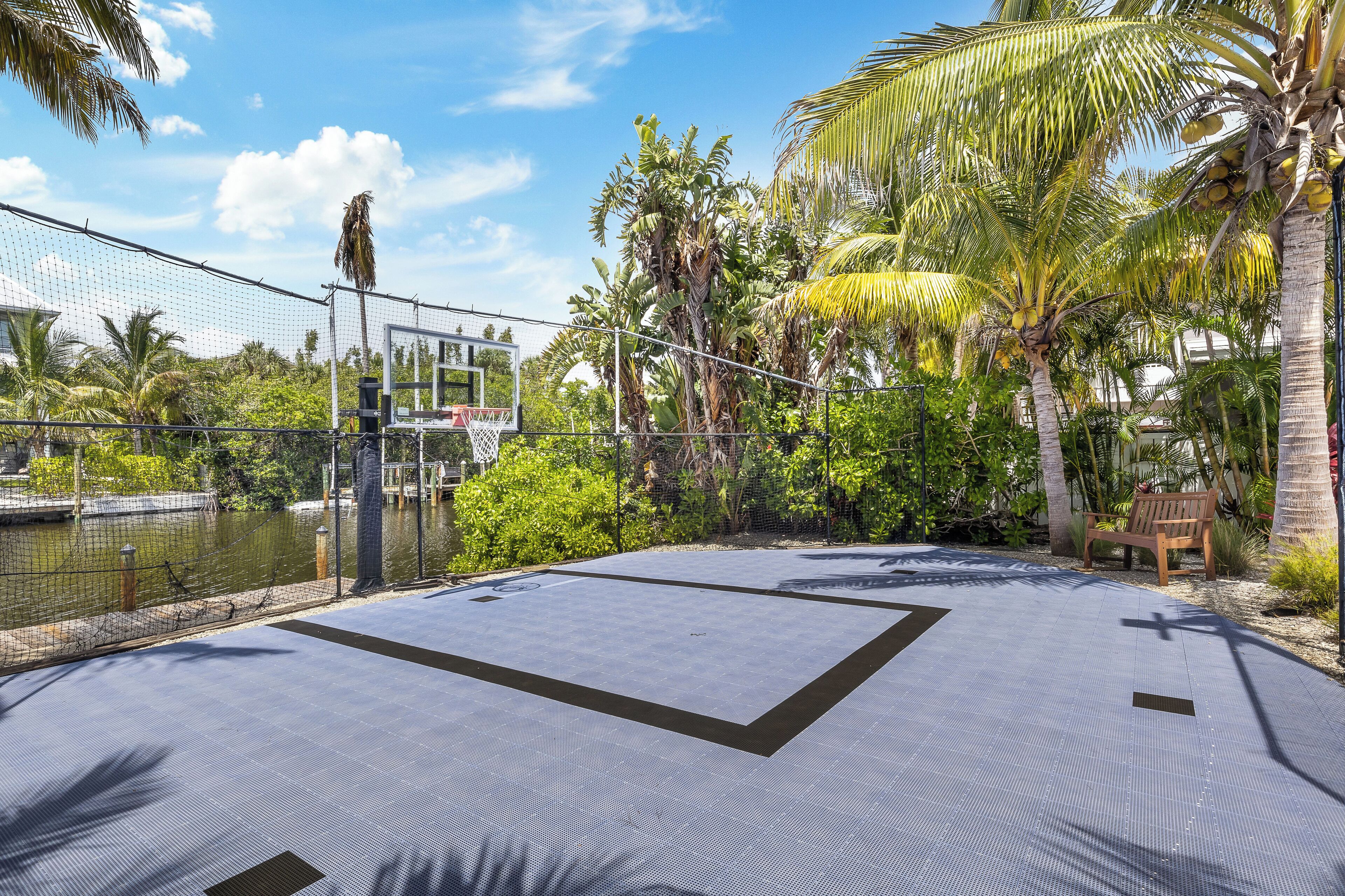 An outdoor basketball court with a net, surrounded by palm trees and a fence.