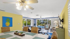 Un coin repas avec une table dressée pour quatre personnes, entouré de chaises en osier, d’un ventilateur de plafond et offrant une vue sur une piscine et un espace de détente extérieur.