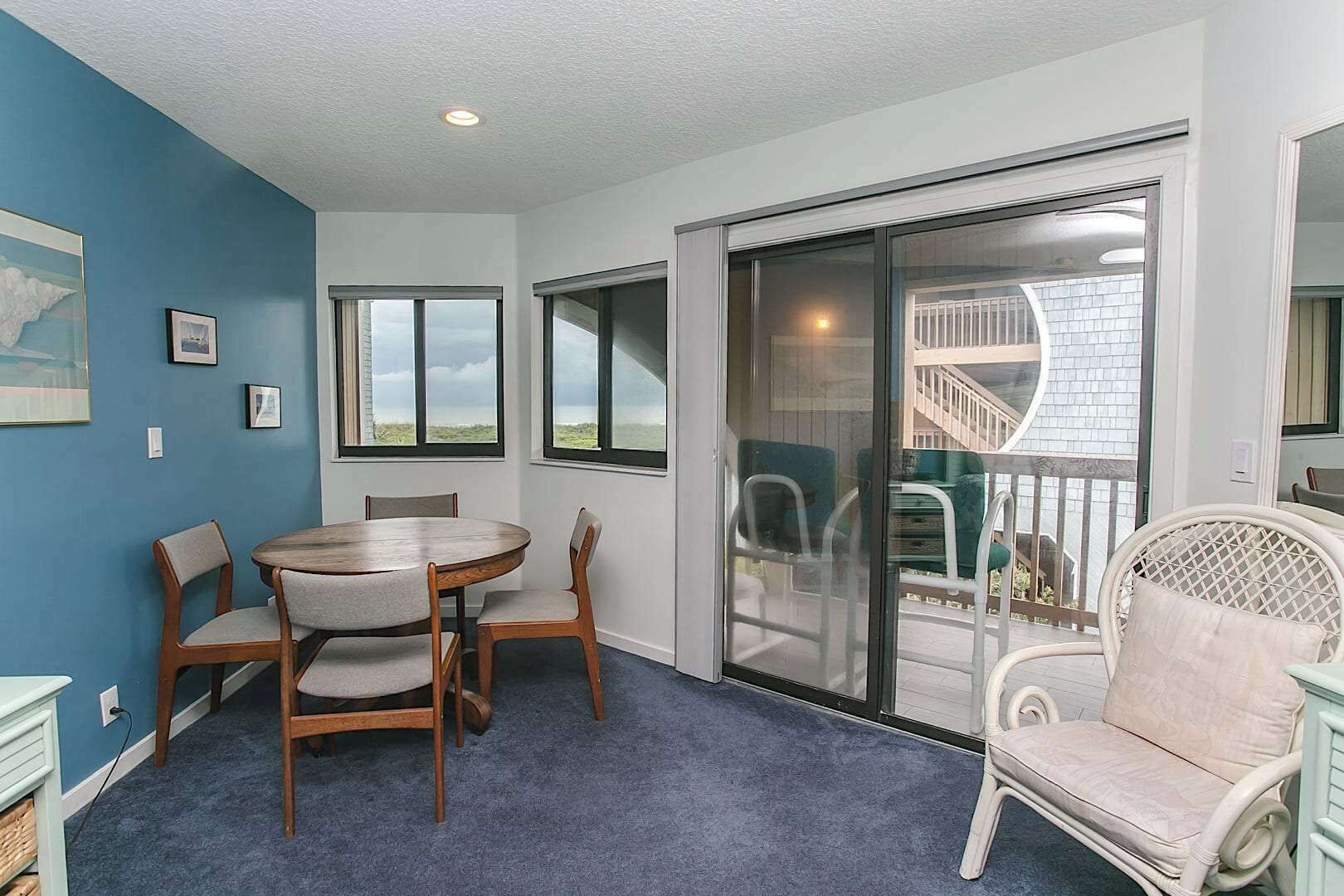 Una habitació amb una taula rodona de fusta i cadires, una àmplia porta corredissa de vidre que dona a un balcó, i una paret d'accent blau.