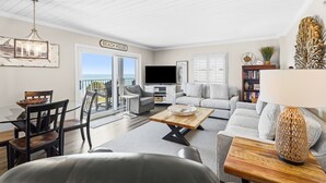 A living room with a dining table, chairs, a sofa, a TV, and a balcony with a sign reading 'Beach House'.