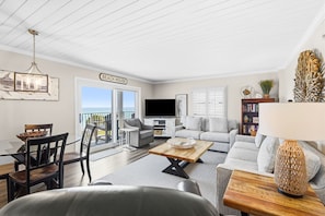 Een woonkamer met een eettafel, stoelen, een bank, een televisie en een balkon met een bordje waarop staat 'Beach House'.