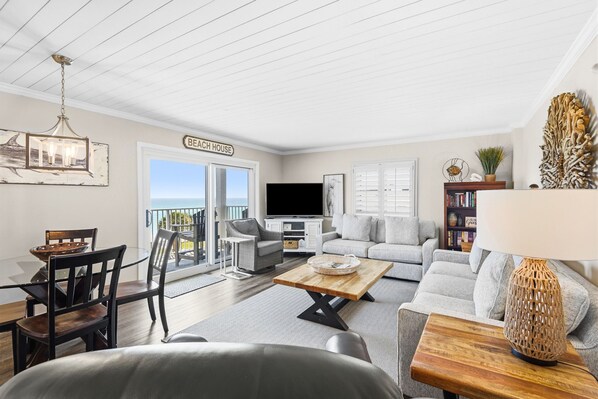 A living room with a dining table, chairs, a sofa, a TV, and a balcony with a sign reading 'Beach House'.
