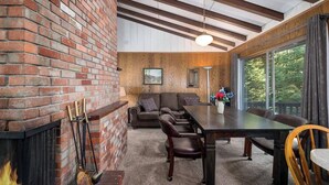 Una sala de estar acogedora con chimenea, un sofá, una mesa de comedor y vistas amplias a través de grandes ventanales.