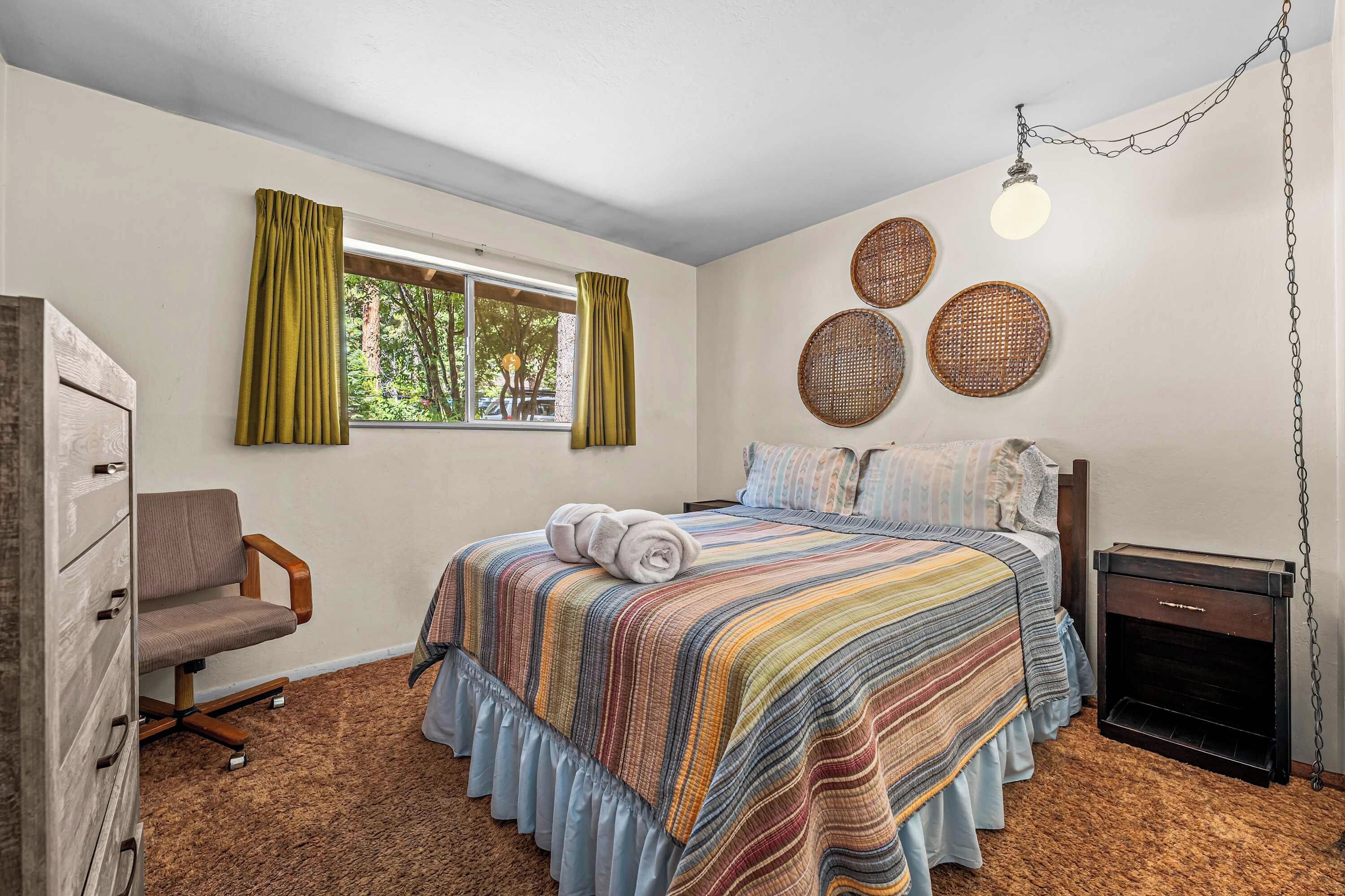 A bedroom with a bed, a chair, a dresser, a window with curtains, and a wall with three woven baskets.