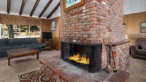 Una sala de estar con chimenea de ladrillo, un sofá, un televisor y una ventana con vistas a árboles.