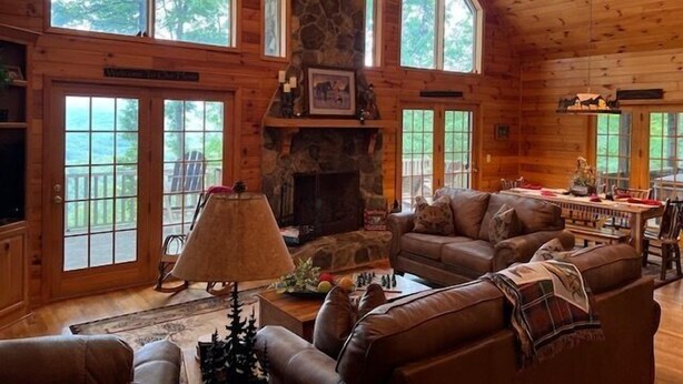 A spacious log cabin interior with a stone fireplace, leather furniture, and large windows offering a view of the surrounding forest. - Sunset Ridge - New Hot Tub! Log Cabin, Views, Deck, Trails (Ferguson)