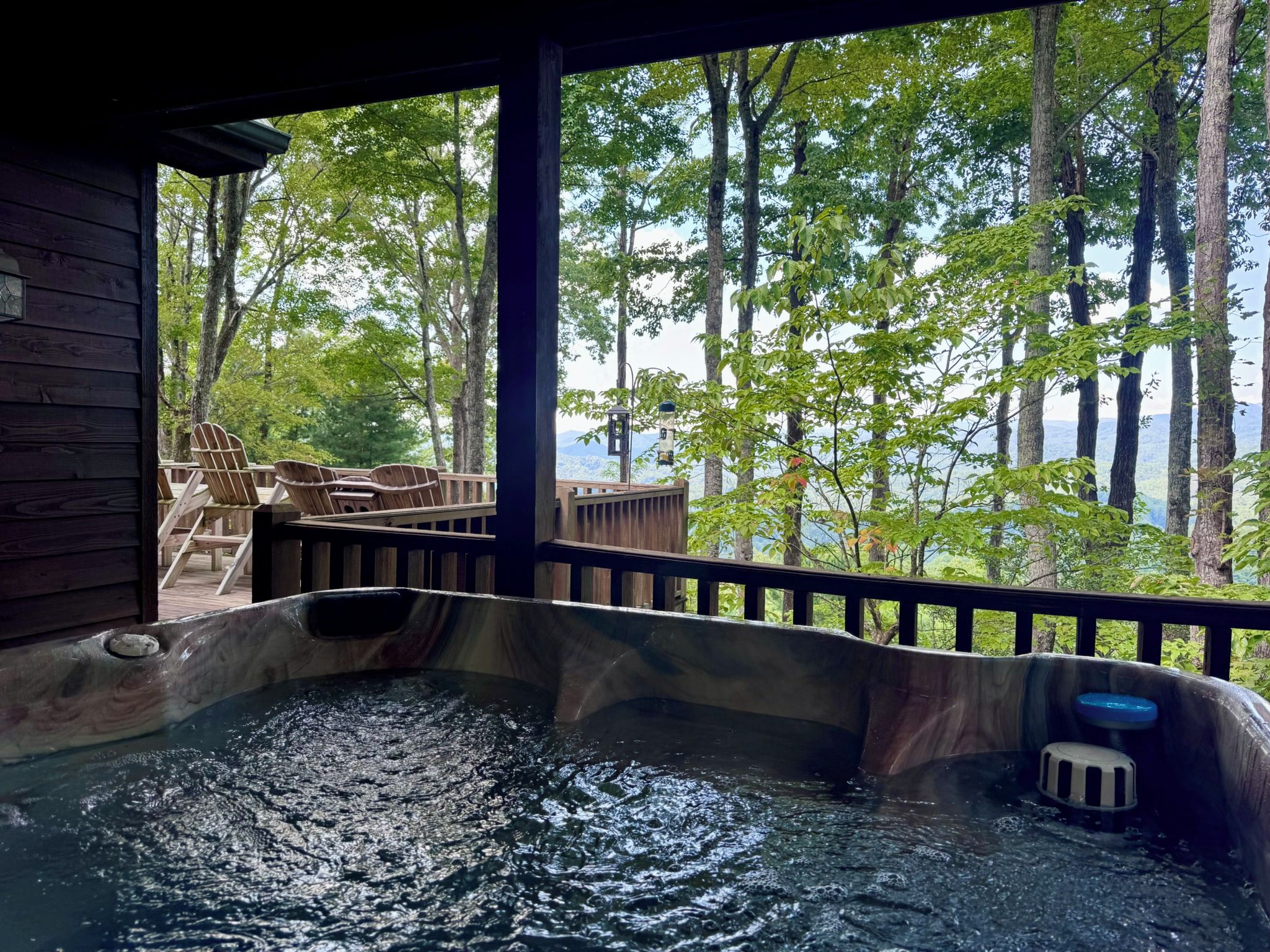 A hot tub with a wooden deck and chairs, surrounded by trees and a view of a lake.