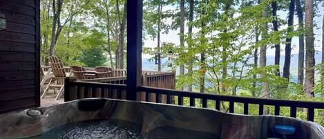 A hot tub with a wooden deck and chairs, surrounded by trees and a view of a lake.