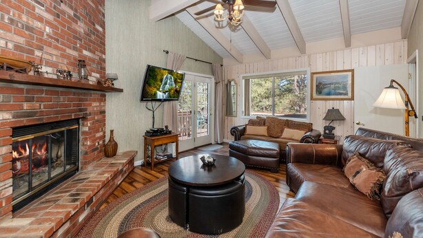 A cozy living room with a brick fireplace, leather sofas, a ceiling fan, and a television mounted on the wall. - Whispering Bear Lodge by Destination Big Bear : Pool Table! Foosball! Views! Internet! Deck! (Big Bear City)
