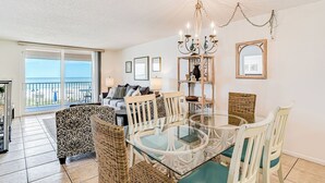 A dining area with a glass table surrounded by wicker chairs, a chandelier, and a view of the ocean through large windows.