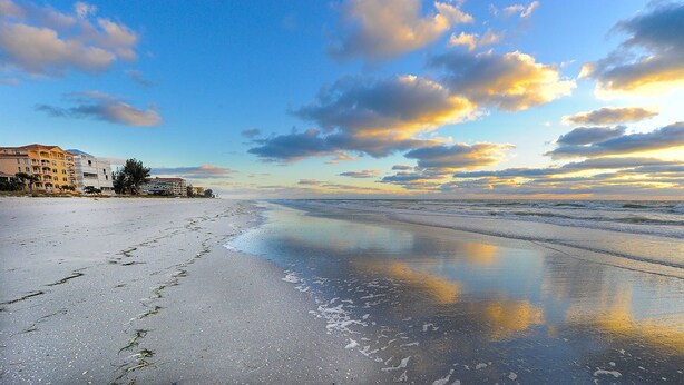 A beach with a clear sky, a calm sea, and buildings in the background.