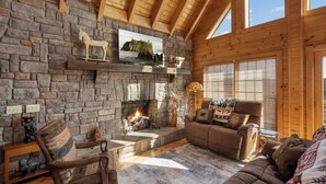 A cozy cabin interior with a stone fireplace, wooden beams, a leather sofa, and a large window.
