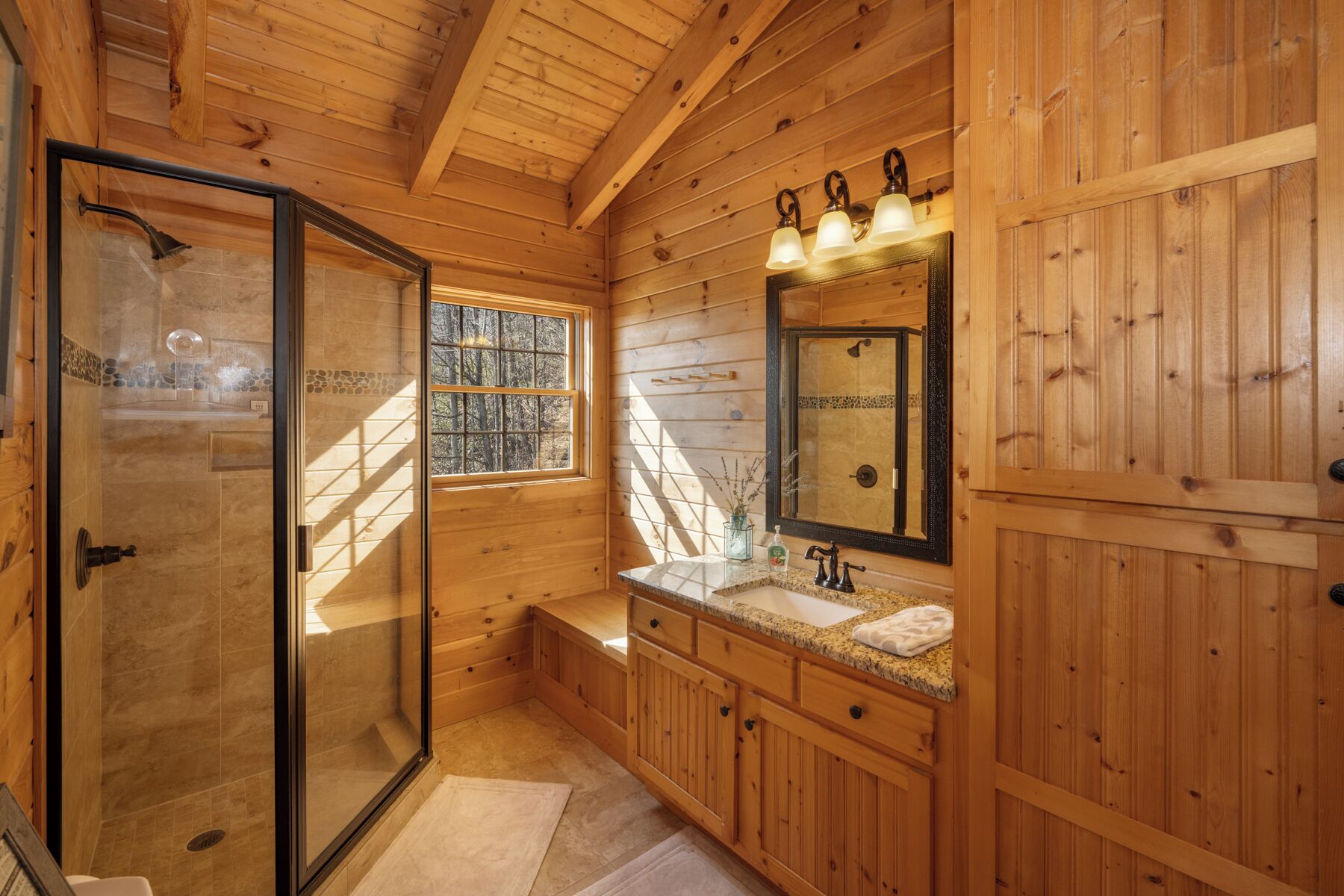 A wooden cabin bathroom with a glass-enclosed shower, a vanity with a sink, and a large mirror with three lights.