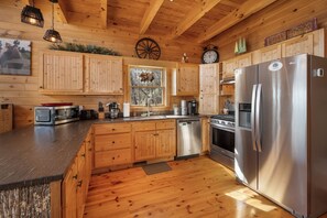A wooden cabin kitchen with a stainless steel refrigerator, a stove, and a dishwasher. There is a window with a view of trees and a clock on the wall.