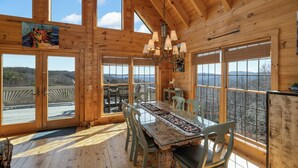 A wooden cabin with a dining area featuring a glass table and chairs, large windows offering scenic views, and a chandelier.