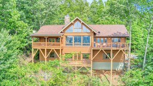 Une cabane en rondins à deux étages, dotée d’un balcon et de grandes fenêtres, entourée d’une forêt dense.