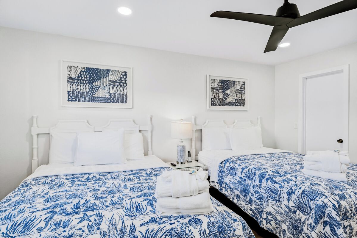 Habitación de hotel con dos camas, cabeceros blancos, colchas de flores azules, toallas blancas, una mesita de noche con lámpara y ventiladores de techo.