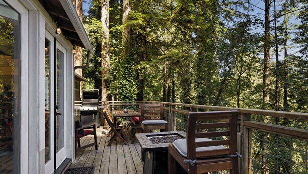 Una terraza de madera con mobiliario exterior, una parrilla y vistas a un área boscosa.