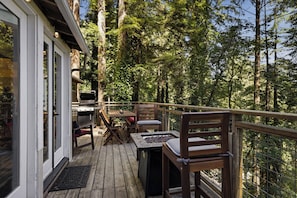 Una terraza de madera con mobiliario exterior, una parrilla y vistas a un área boscosa.