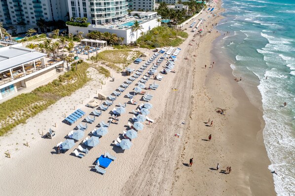 An aerial view of a beach with rows of sun loungers and umbrellas, a pool, and a hotel building in the background.