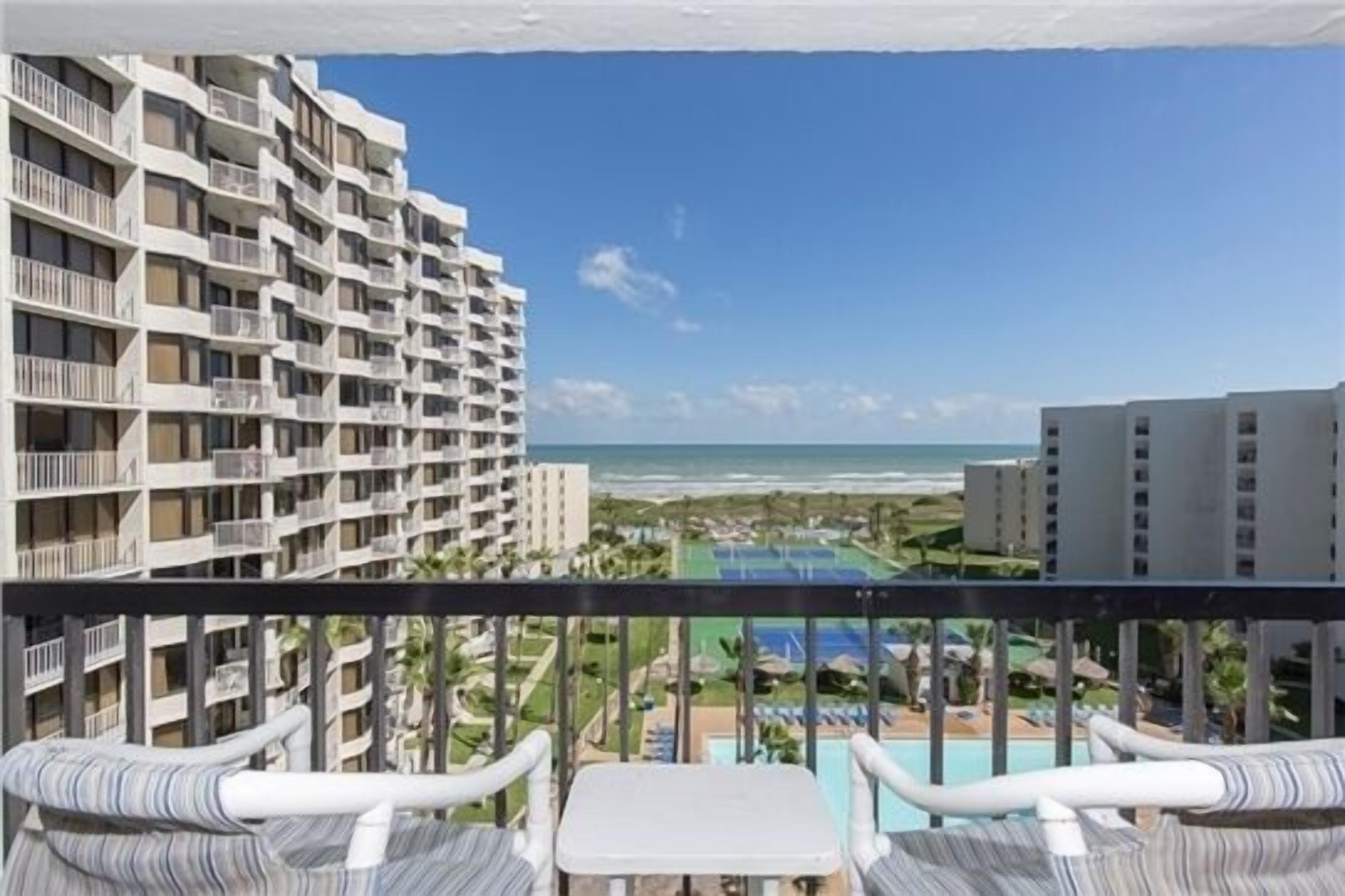 A balcony with white wicker furniture overlooking a resort area with a pool and ocean view.