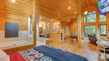 Interior de una cabaña de madera con una cama grande, una mesa de billar, una mesa de futbolín y un televisor montado en la pared.