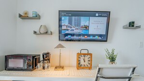 Quarto de hotel com televisão de tela plana exibindo uma mensagem de boas-vindas, cafeteira, relógio e escrivaninha com cadeira.