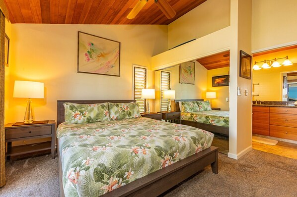 Une chambre avec un grand lit, deux tables de chevet, une commode, un miroir et une salle de bain équipée d’un lavabo et d’un meuble-lavabo.