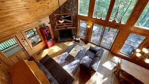 Ein gemütliches Blockhaus-Interieur mit Kamin aus Naturstein, Holzwänden und großen Fenstern mit Blick auf den Wald.