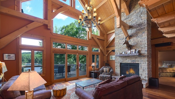 A spacious living room with a stone fireplace, leather sofas, and a large window overlooking a deck. - Elegant Lakefront Retreat w/ Hot Tub & Fireplaces (Oakland)