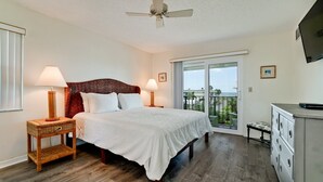 A bedroom with a bed, two bedside lamps, a ceiling fan, a balcony with a view, and a dresser.