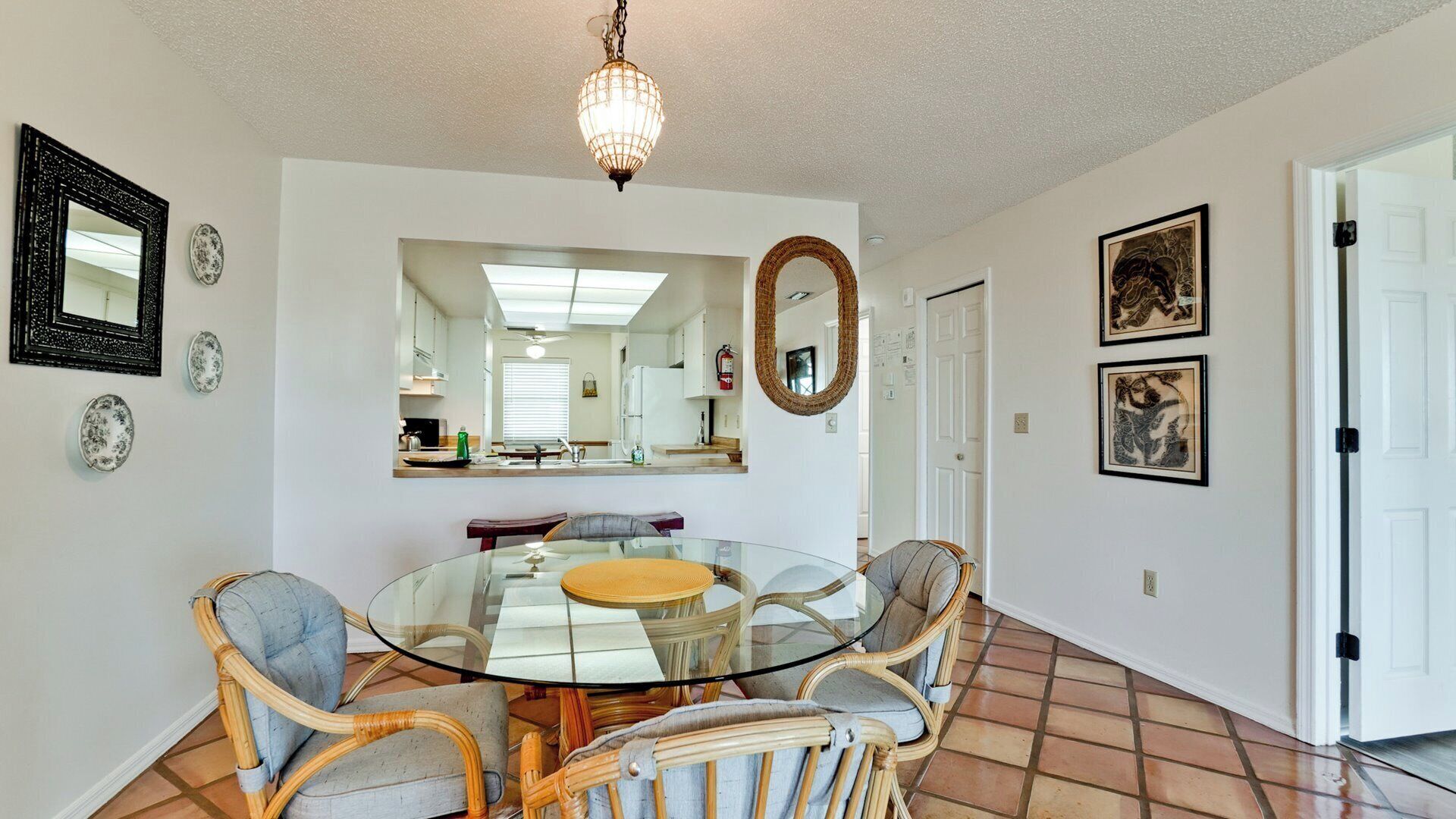 A dining area with a glass table surrounded by wicker chairs, a hanging light fixture, and a kitchen visible through a glass partition.