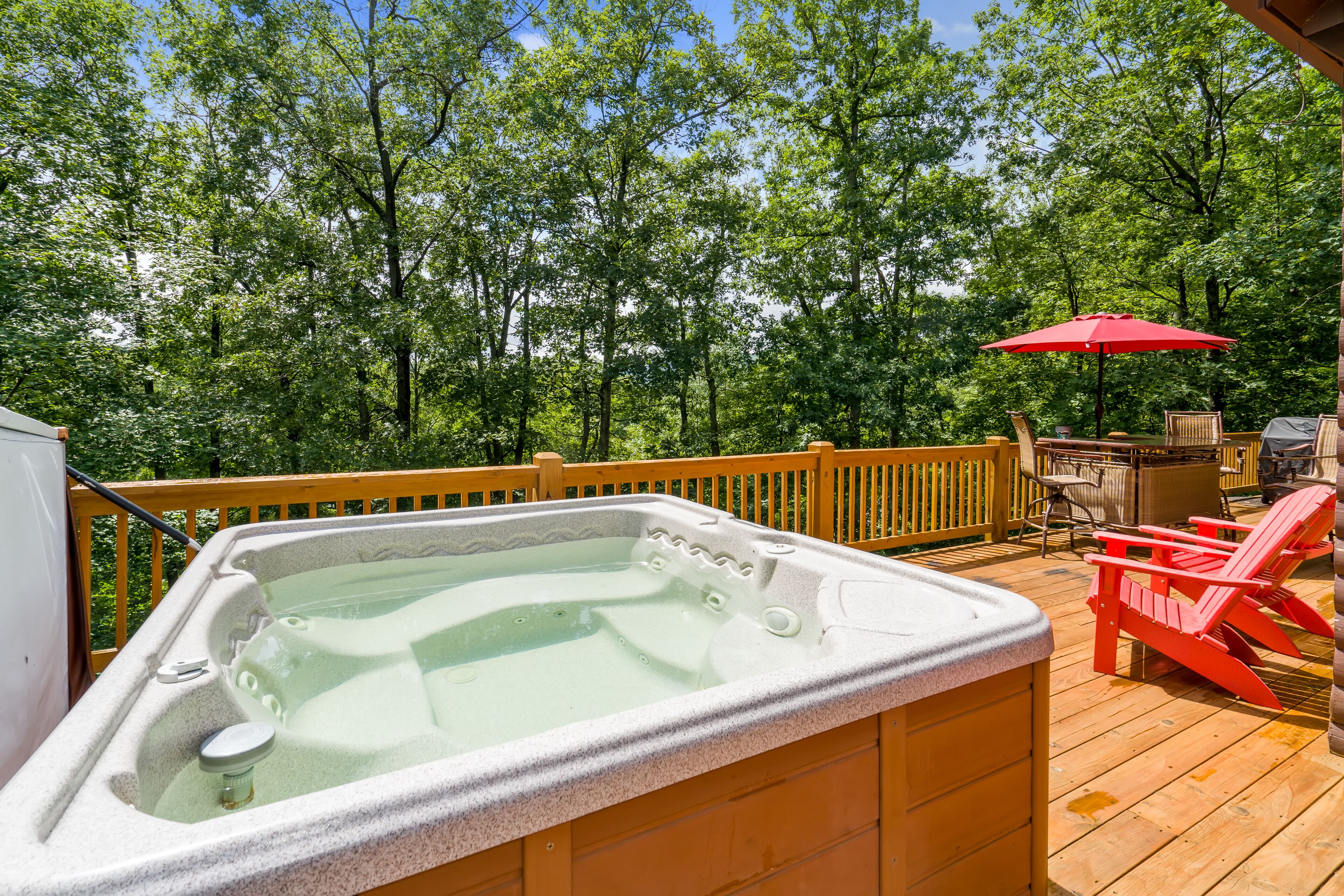 A hot tub on a wooden deck with a view of trees, a red umbrella, and outdoor furniture.